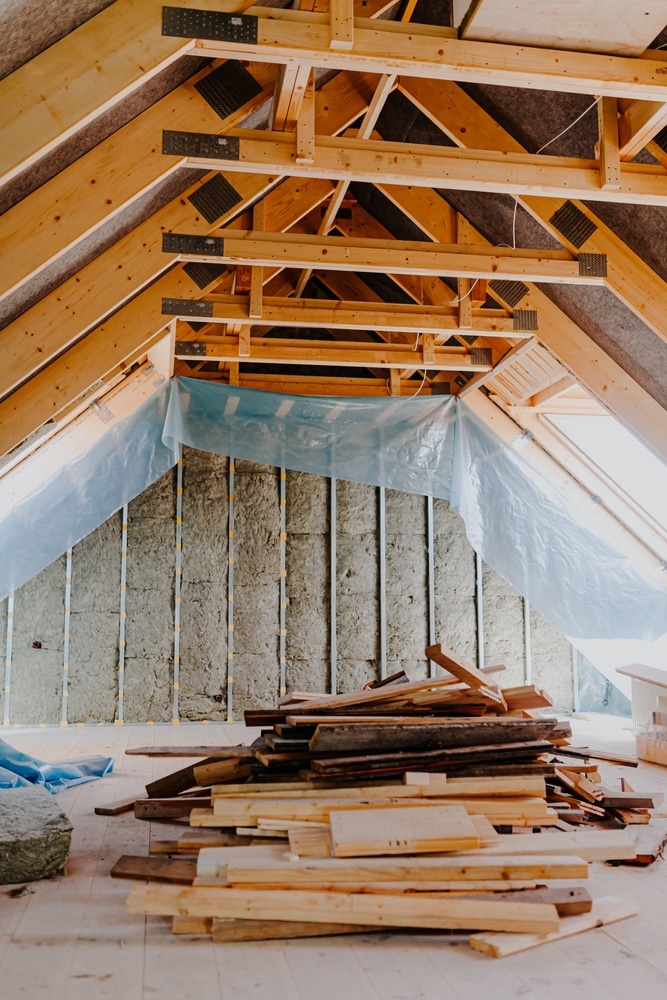 Charpente bois et isolation : travaux de rénovation de combles. Intérieur de combles en rénovation. Charpente en bois apparente, isolation posée entre les montants, bâche plastique et pile de bois au sol.