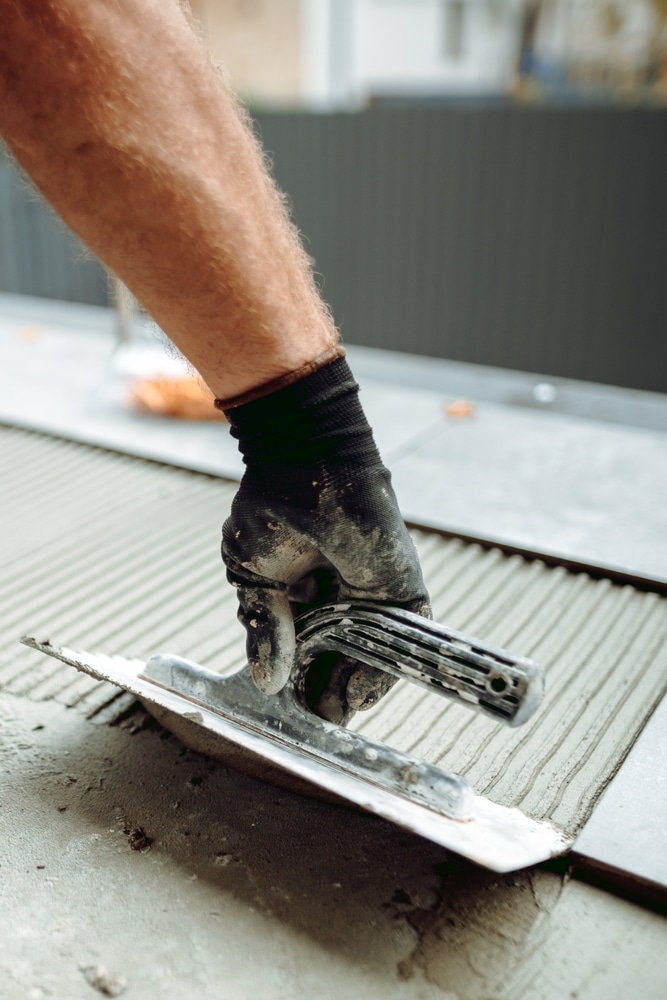 Pose Carrelage : étalez le ciment colle avec une truelle crantée Artisan utilisant une truelle crantée pour étaler du mortier colle sur un sol en béton, préparant la pose de carrelage. Gant noir.