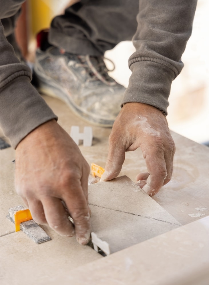 Pose de carrelage : les mains expertes du carreleur Mains d'un carreleur professionnel ajustant un carreau en céramique avec des clips de nivellement. Chaussure de sécurité en arrière-plan.