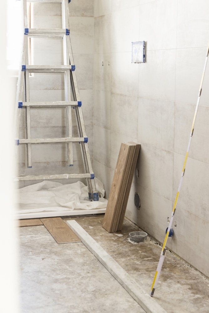 Chantier de rénovation : carrelage mural et matériaux prêts. Rénovation de salle de bain : mur carrelé beige, échelle en aluminium et planches de bois sur sol béton. Outils de mesure et matériaux.