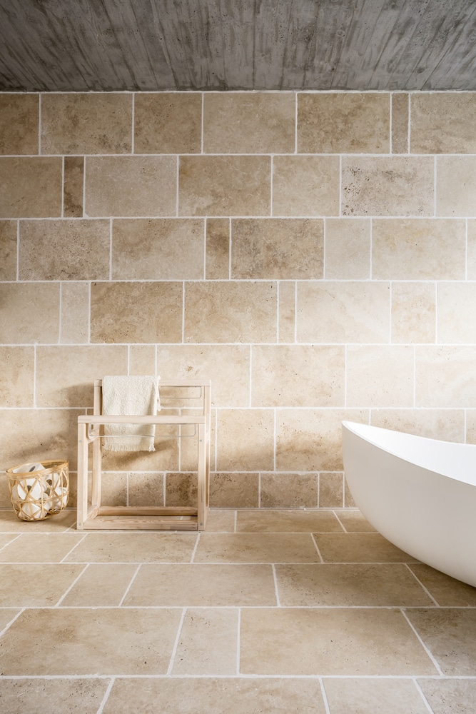 Travertin beige : l'élégance naturelle pour votre salle de bain Salle de bain minimaliste. Murs et sol en dalles de travertin beige. Baignoire îlot blanche, porte-serviettes en bois et plafond béton.