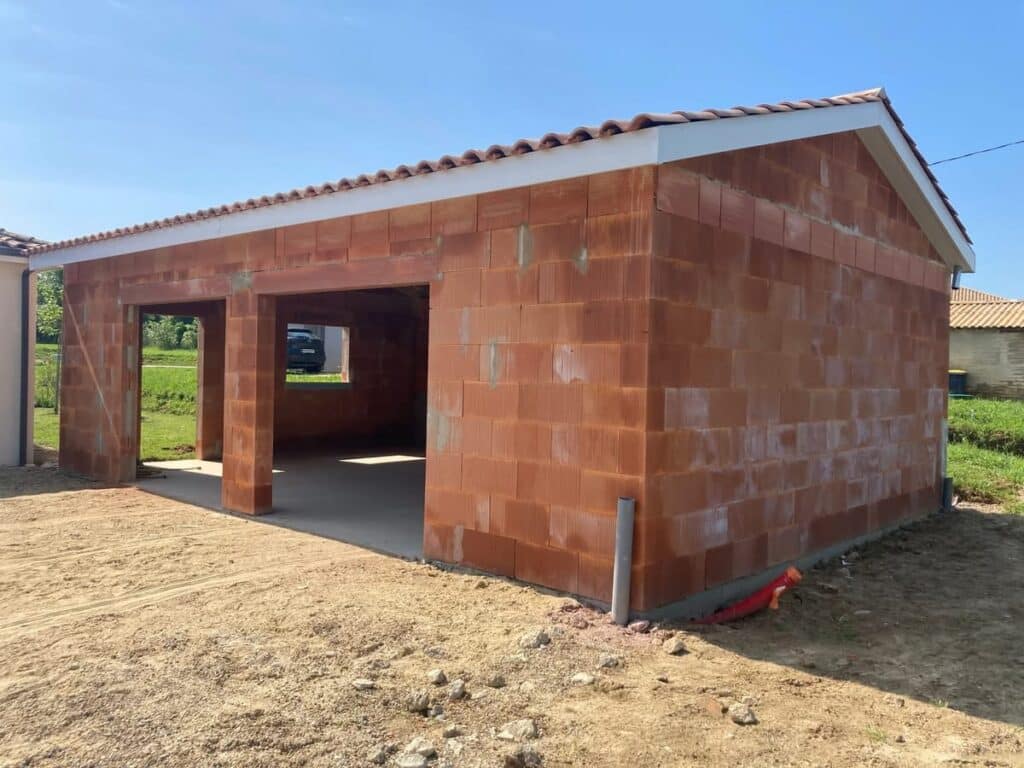Gros œuvre terminé : Garage double en briques terre cuite. Gros œuvre d'un garage double en briques monomur rouges, avec toiture. Deux grandes ouvertures et sol en dalle béton sur chantier.