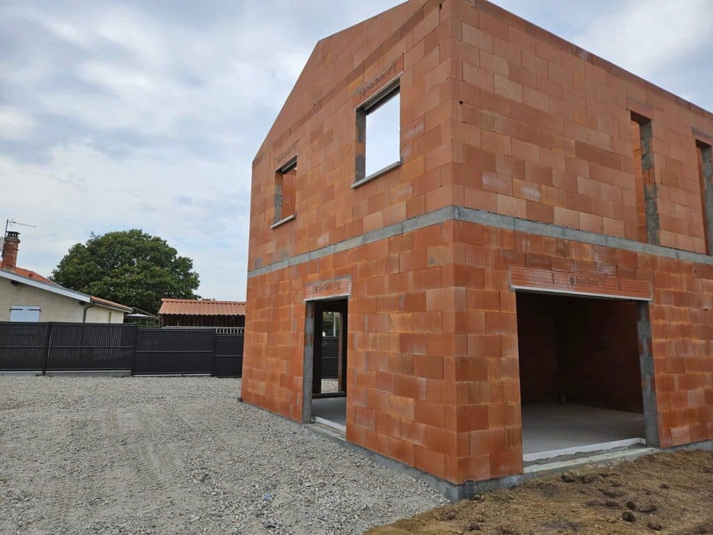 Construction de maison individuelle. Façade en briques terre cuite "bio'bric" sous un ciel nuageux. Gros œuvre achevé avec portes et fenêtres.