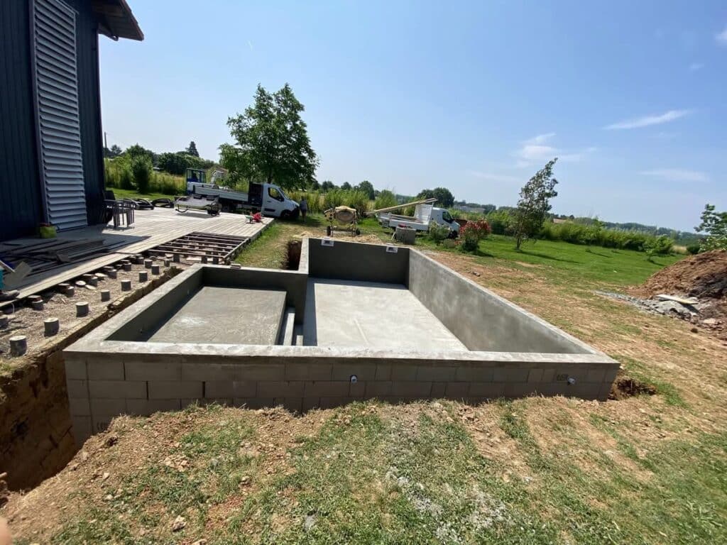 Construction piscine béton, structure et terrasse en cours. Structure de piscine rectangulaire en béton avec zone peu profonde (plage), construction avancée, adjacente à la fondation d'une terrasse en bois.