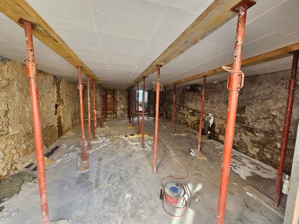 Rénovation : étaiement essentiel d'une dalle en sous-sol. Étaiement de sous-sol. Longue pièce en rénovation avec étais rouges, poutres en bois, plafond blanc et murs de pierre bruts.