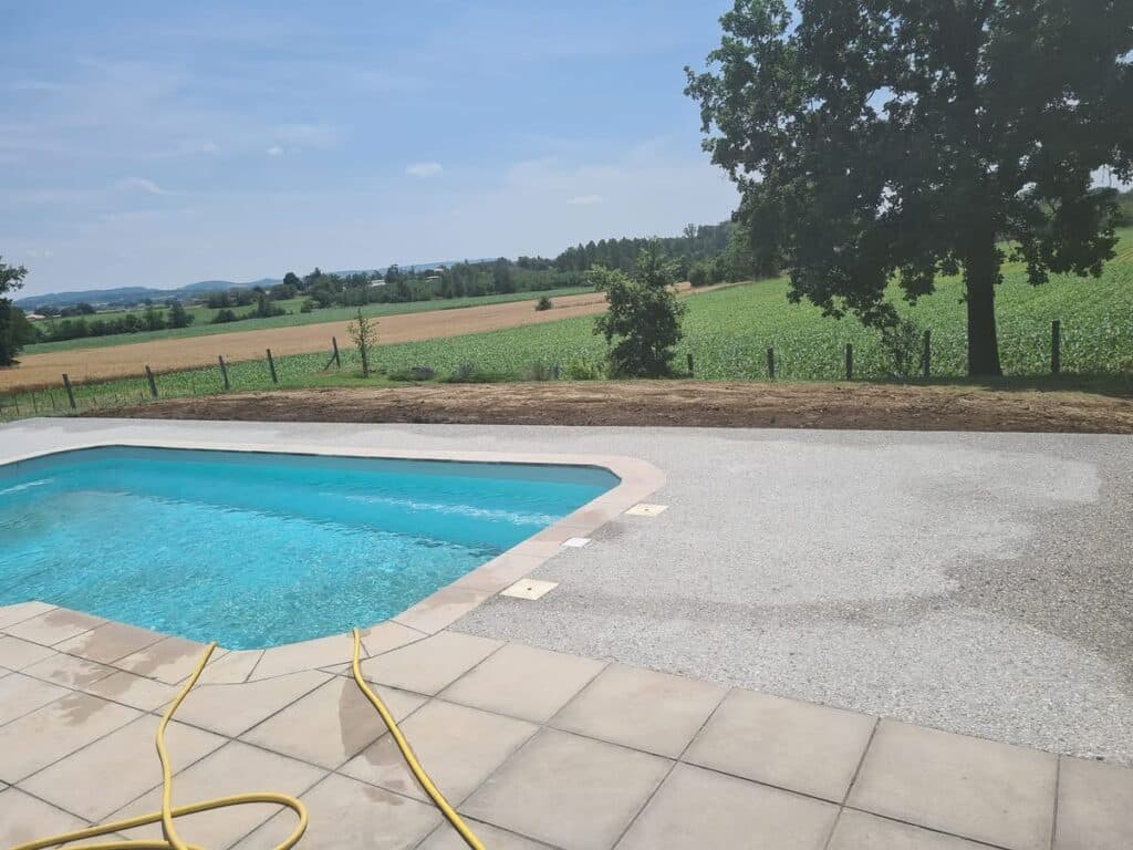 Piscine et terrasse en agrégats : vue imprenable sur la campagne. Piscine extérieure avec margelles beiges et terrasse en gravier agrégat. Vue panoramique sur champs agricoles et ciel bleu sous un grand chêne.